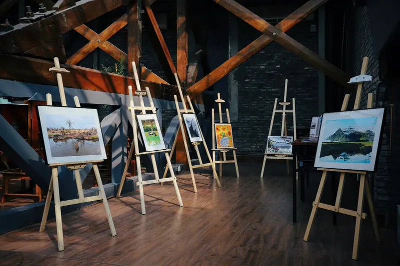 Houten schildersezels met ingelijste schilderijen en tekeningen in een lichte atelierruimte met oude houten balken.